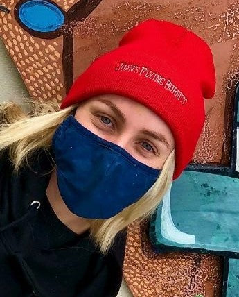 A masked woman wearing a red beanie with "Juan's Flying Burrito" embroidered across the cuff
